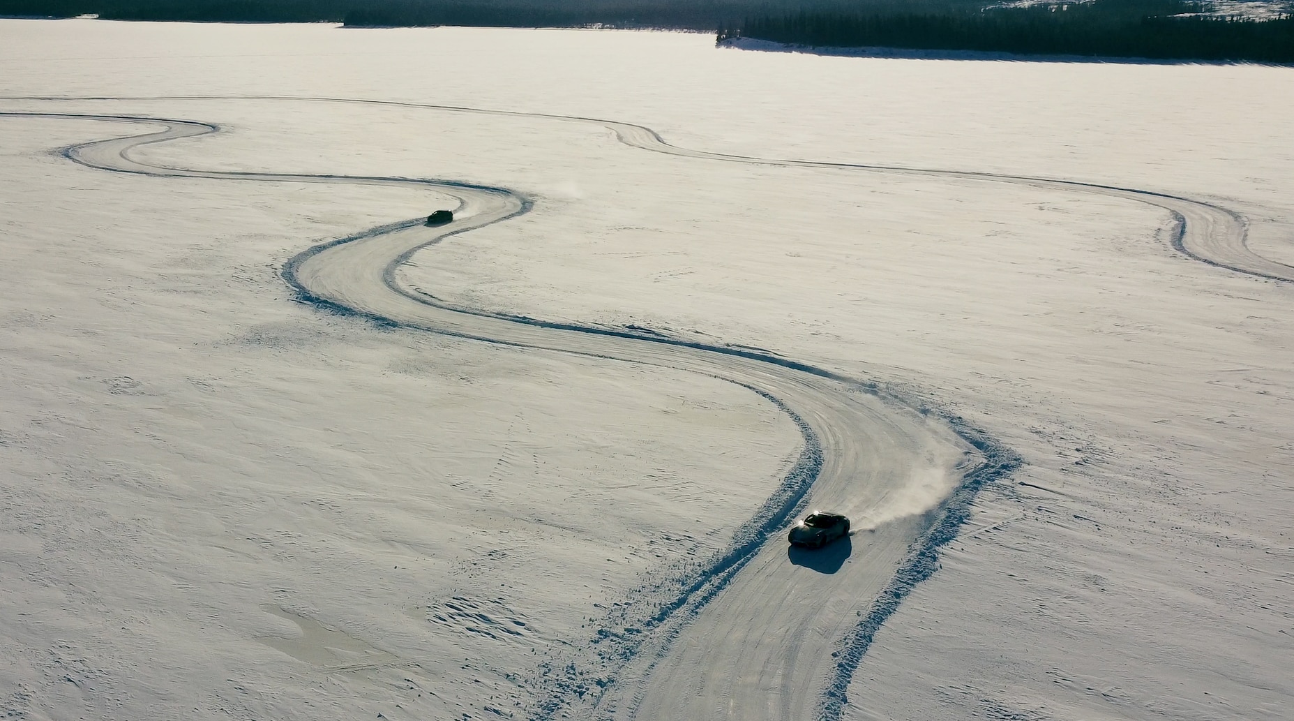 Birds eye view of two cars driving on a snowy car track on a frozen lake.