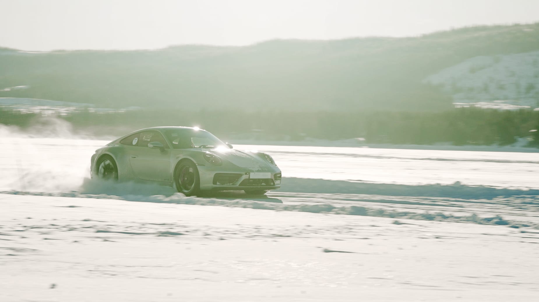 A silver Porsche 911 Carrera GT4s driving along a snowy path in an open environment